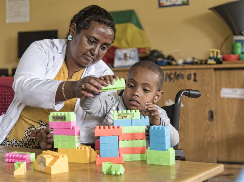 A teacher builds with colourful blocks alongside a young boy who uses a wheelchair.