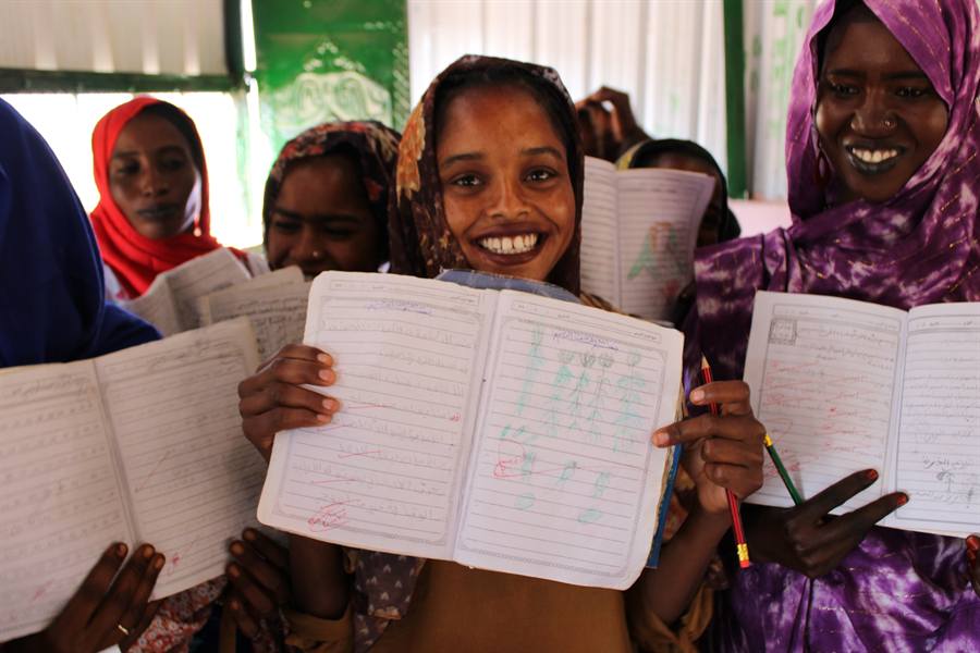 Young women in a displacement affected community proudly hold open their exercise books to show pages of completed literacy work.