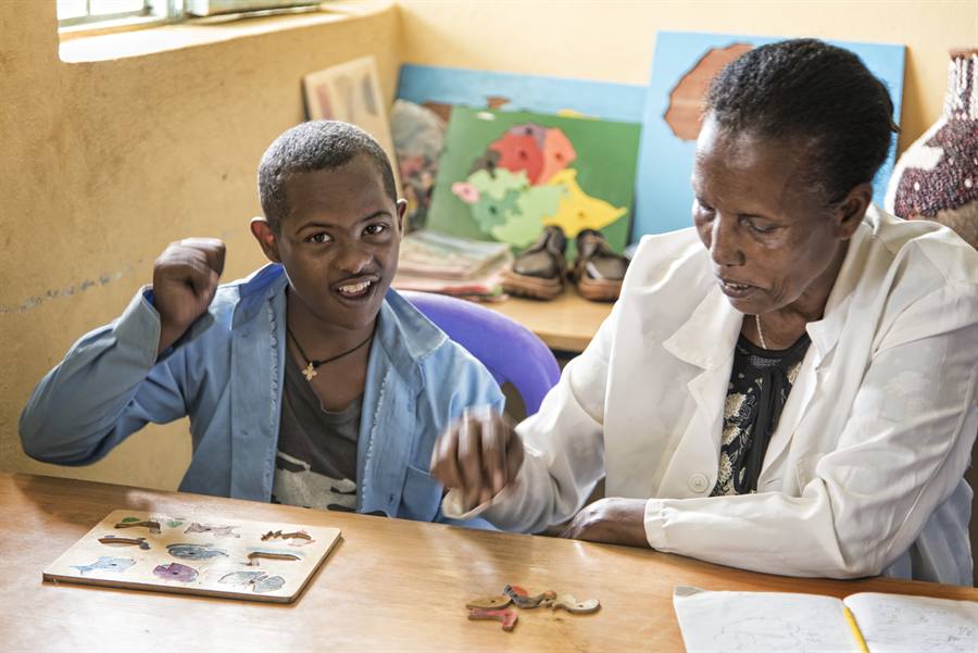 A young learner celebrates completing a wooden animal puzzle beside his teacher, with a map of Africa and Ethiopia puzzle boards displayed behind them.