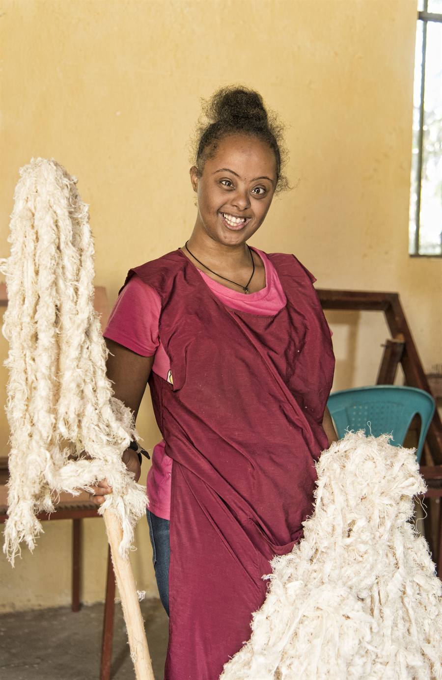 A young woman with Down syndrome smiles proudly while holding hanks of spun cotton in a vocational weaving workshop.